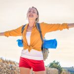 Happy woman basking in sunlight on a summer hike with yoga mat.